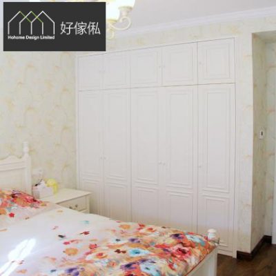 睡房一幅乳白色古典風入牆衣櫃，連上方一排掩門吊櫃，門板有方形線框浮雕；牆身貼淺黃花卉牆紙，前方有白色床頭同床頭櫃，床上鋪彩色花卉床品，左上角有好傢俬 Hohome 標誌。