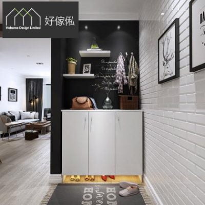 玄關近景，白色三門鞋櫃鑲入黑色凹陷牆位，櫃底留空藏燈帶照亮地面高踭鞋，上方掛黑色掛勾層板放綠植，右邊白磚牆掛鹿頭線條裝飾畫，地下鋪字母圖案地氈放拖鞋，左側望向客廳。