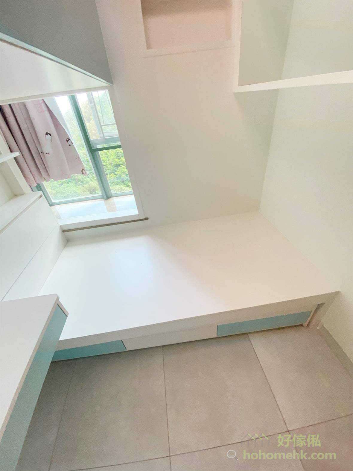 訂造地台床貼住窗邊，白色床板配淺藍色床下抽屜面板，左側有一張同色矮枱及鏡面，窗外見綠樹配粉紅色窗簾，右上方有白色開放層架，地板鋪淺灰色磁磚，整體淡綠白配薄荷藍配色。