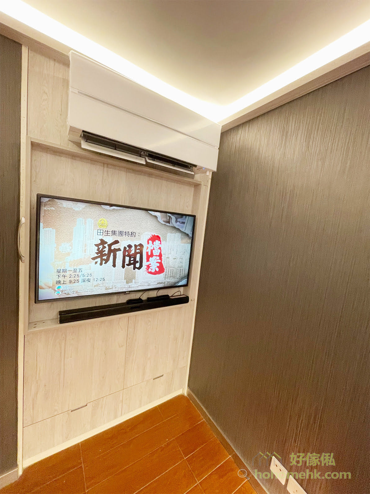 睡房電視櫃使用中，電視播放電視節目，掛喺淺木紋背板上，下方黑色soundbar，上方白色分體冷氣連暖白燈槽天花，右邊深灰木紋牆紙，地下啡色木地板。
