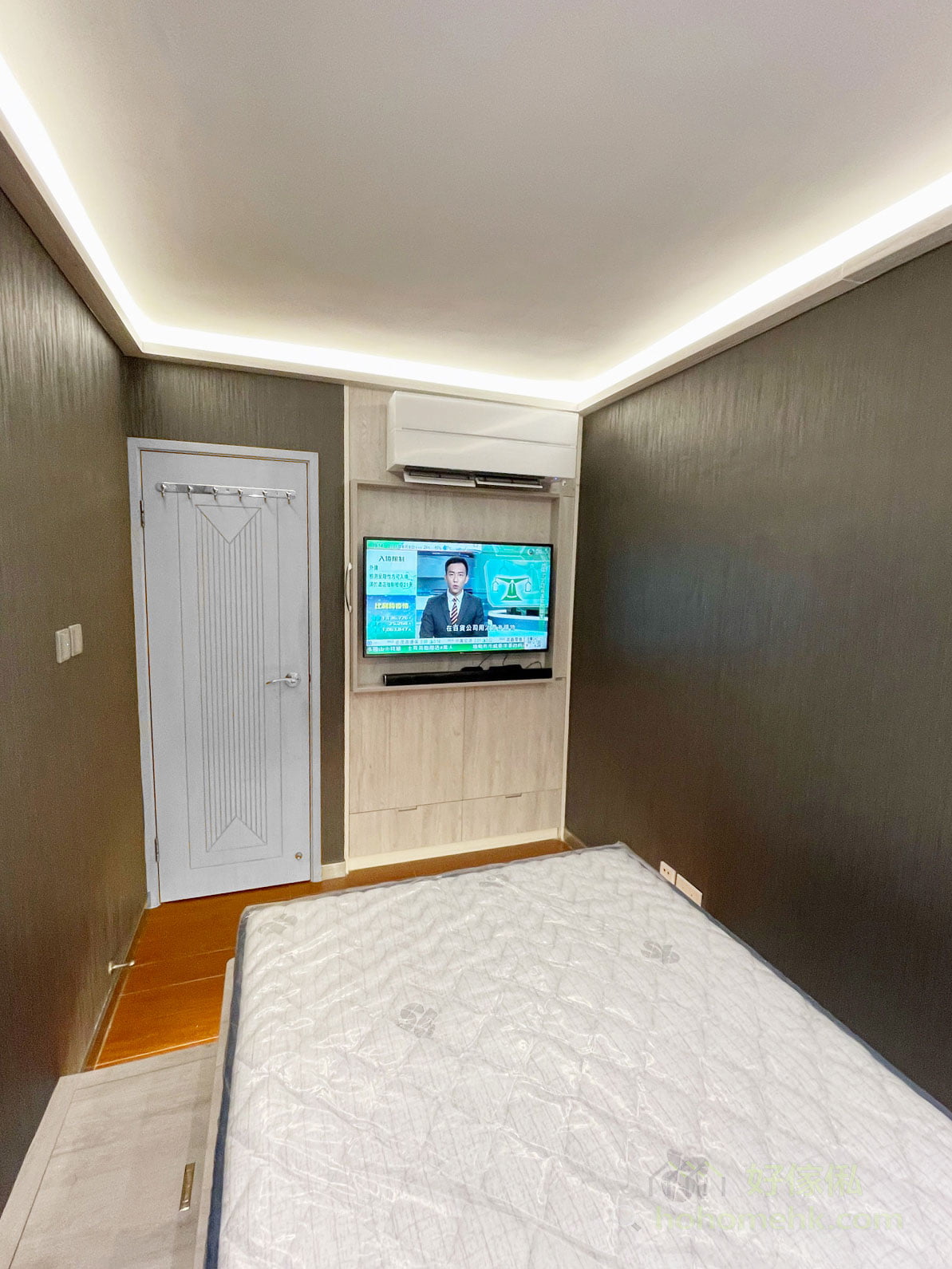 睡房廣角，右邊訂造電視櫃掛住電視連soundbar，淺木紋背板，前方灰色床褥，左邊白灰色木門配掛勾，深灰木紋牆紙，啡色地磚，天花暖白燈槽。