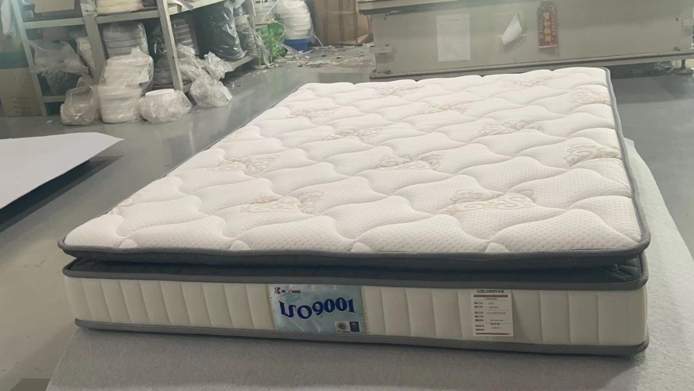 工場內一張床褥平放喺工作枱上，白色布面有弧線格紋同淺色花紋，四周配灰色包邊，奶白色側面有直條車縫及產品標籤，後方可見層架、包裝物料同機器。