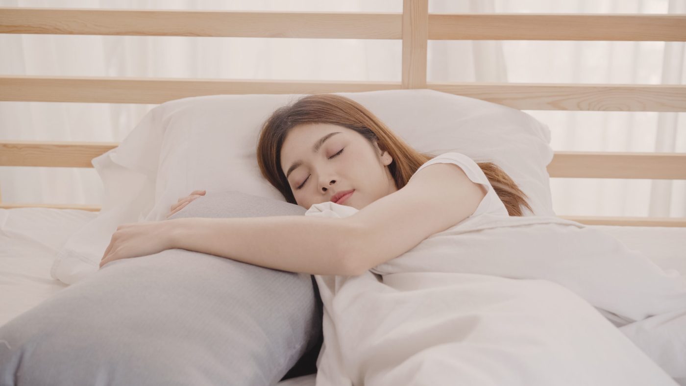 一名長髮女子閉眼側臥喺淺木色上下床嘅上格，攬住一個灰色長形抱枕，鋪白色床單同被舖，背後係淺木橫條床欄同白紗窗簾，畫面光猛柔和。