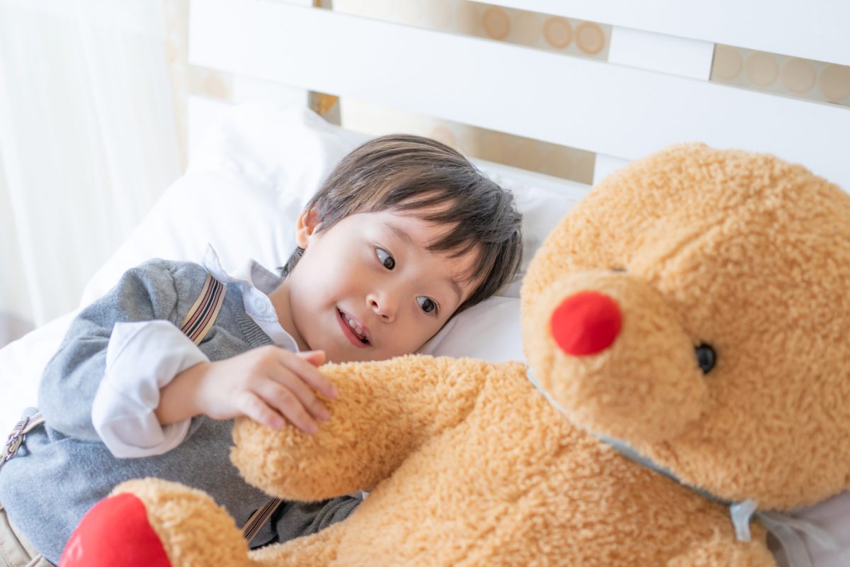 一名短髮男童側臥喺鋪白枕嘅床上,笑住攬住一隻紅鼻啡色大泰迪熊公仔,著灰色背心配白色長袖恤衫,背景係白色橫條床欄同白紗窗簾,畫面光猛溫暖。