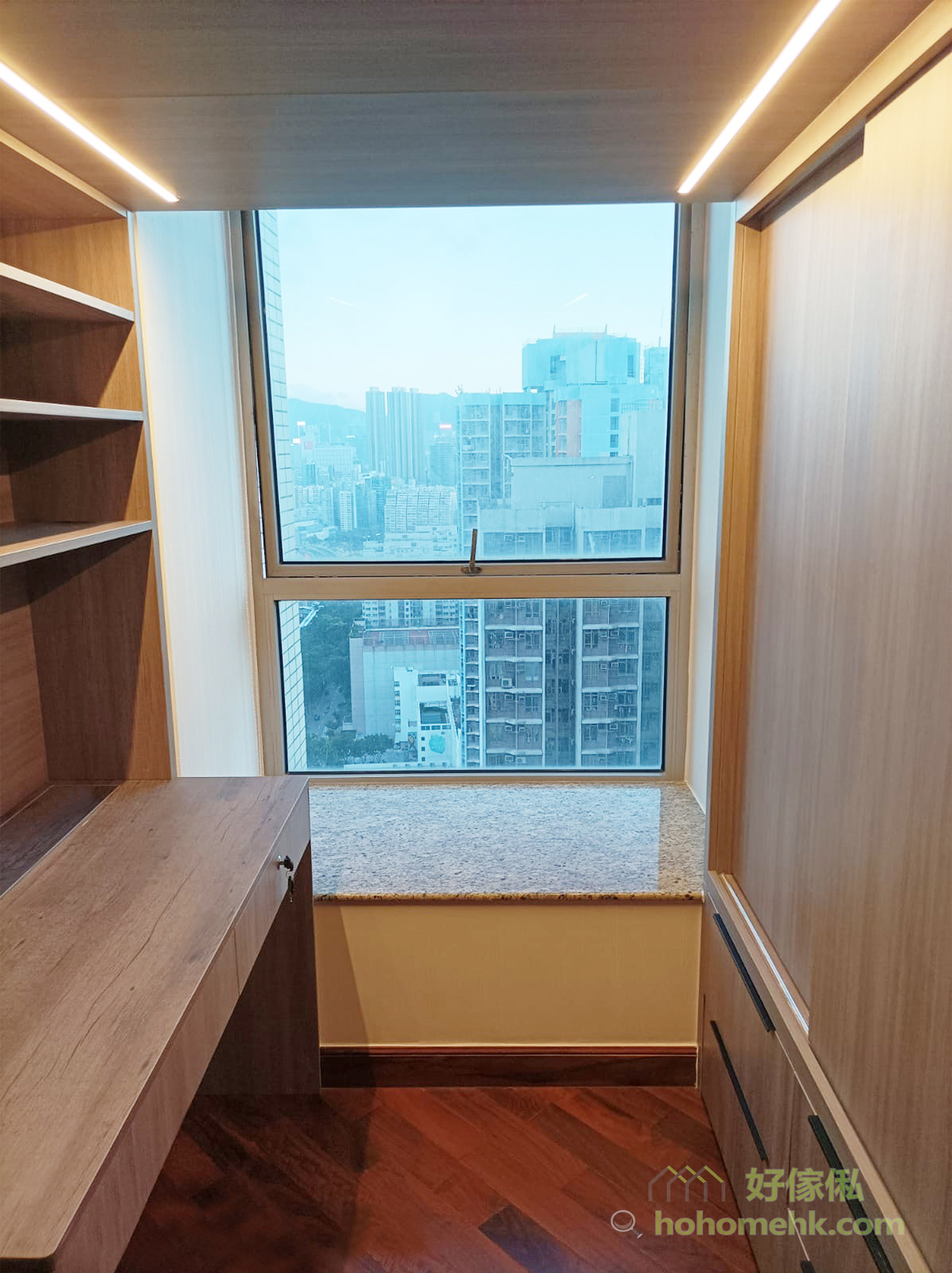 閣樓下層近窗實景，左邊淺木紋書檯連開放層架，中間落地窗下做咗雲石面窗台座位櫃，右邊為淺木紋趟門櫃連抽屜，天花裝LED燈帶，窗外望到密集嘅城市樓宇景，地面為深紅木色地板。