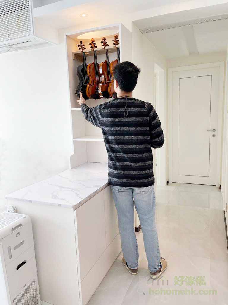 一位穿深灰條紋上衫、淺色牛仔褲的男士背向鏡頭，伸手扶住白色訂造櫃近櫃頂掛架上的小提琴，掛架上排住一個黑色琴盒同幾把棕色小提琴；櫃身純白色，地櫃配白色雲石紋枱面，左側放有白色空氣清新機，右邊係鋪白色地磚嘅走廊同白色房門。