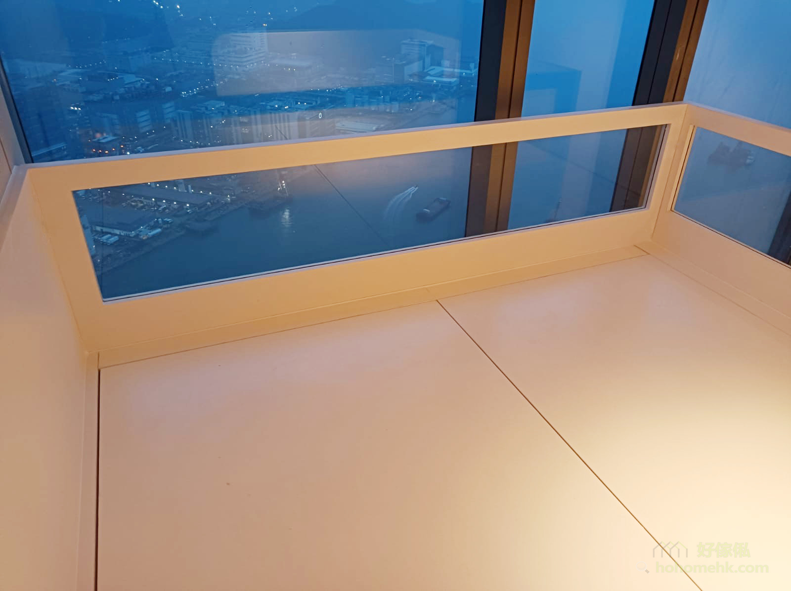 閣樓上層休息平台視角，白色矮牆上方嵌有橫向玻璃圍欄，透過玻璃可見夜間維港城市景色及點點燈光，平台地面以白色板材鋪設，溫暖燈光由下方透出，整體偏暖白色調