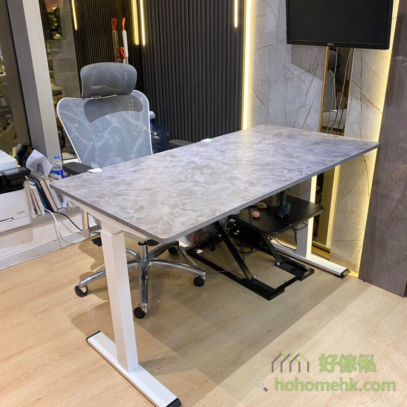 Morton電動升降枱佩茲利限定色全身實拍，深灰雲石裂紋枱面配白色T形升降腳，置於展示空間，枱後設有灰色高背辦公椅，升降腳下可見一部腳踏設備，左側設有牆面直線裝飾條及顯示屏。