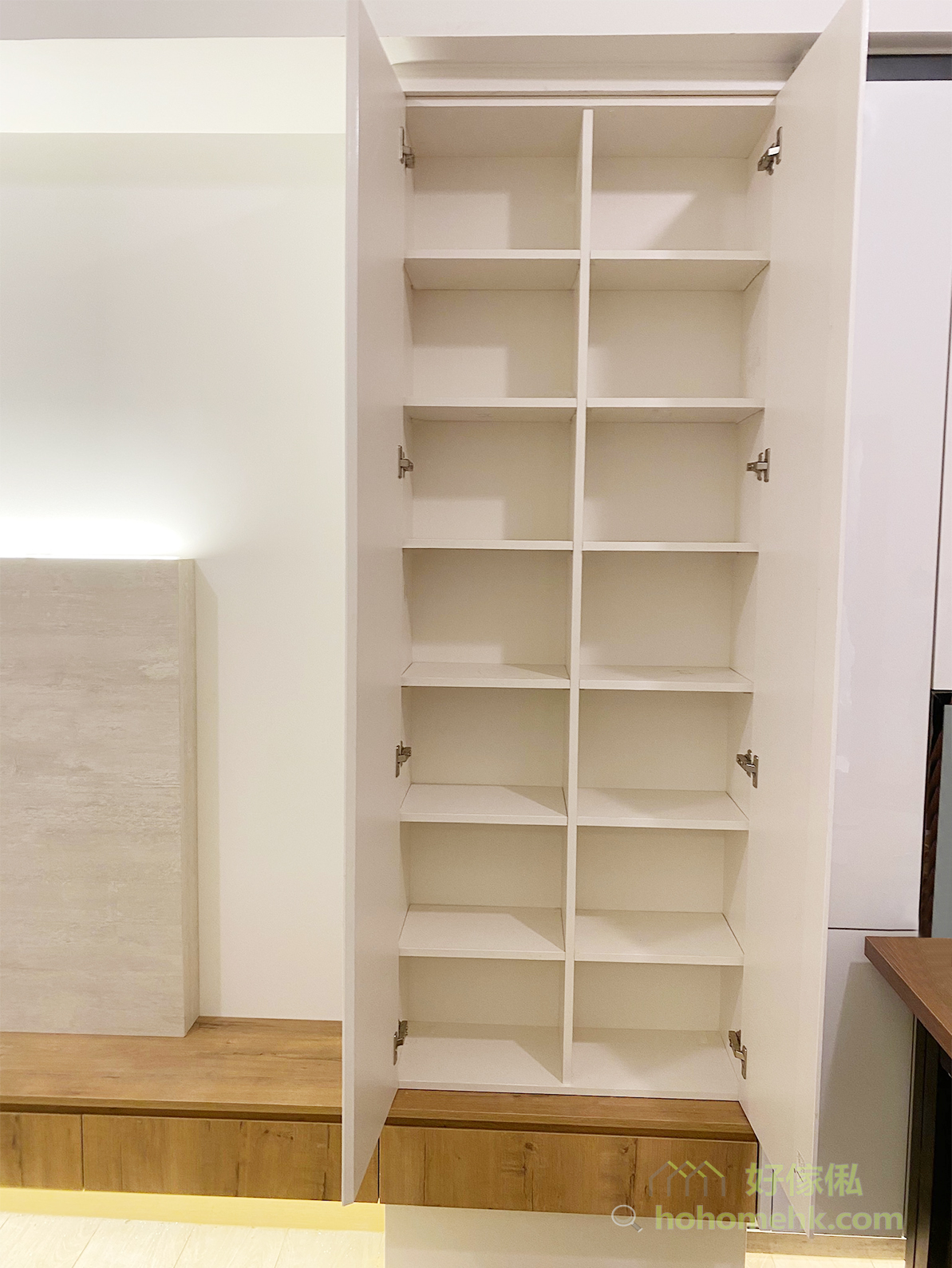 白色到頂儲物櫃開門展示內部：兩扇門板完全開啟，露出左右兩列共約八層白色層板格，隔板間距均勻可按需調整，五金合頁清晰可見；背景可見電視背景板及橡木色地櫃，地板為淺色磁磚