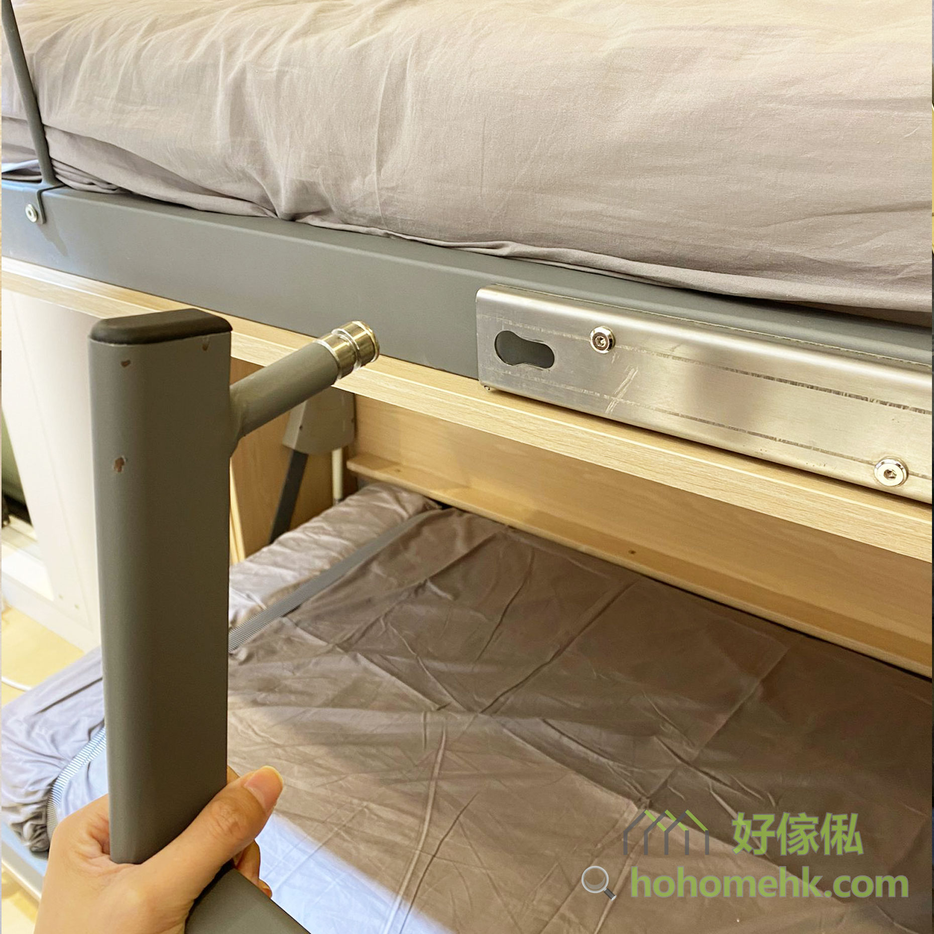 手持灰色掛梯頂端榫扣插入上格床灰色鋁合金側軌橢圓形卡槽，特寫顯示不鏽鋼定位螺絲及插槽位置，上方可見灰色床褥邊，背景為淺橡木色夾板。