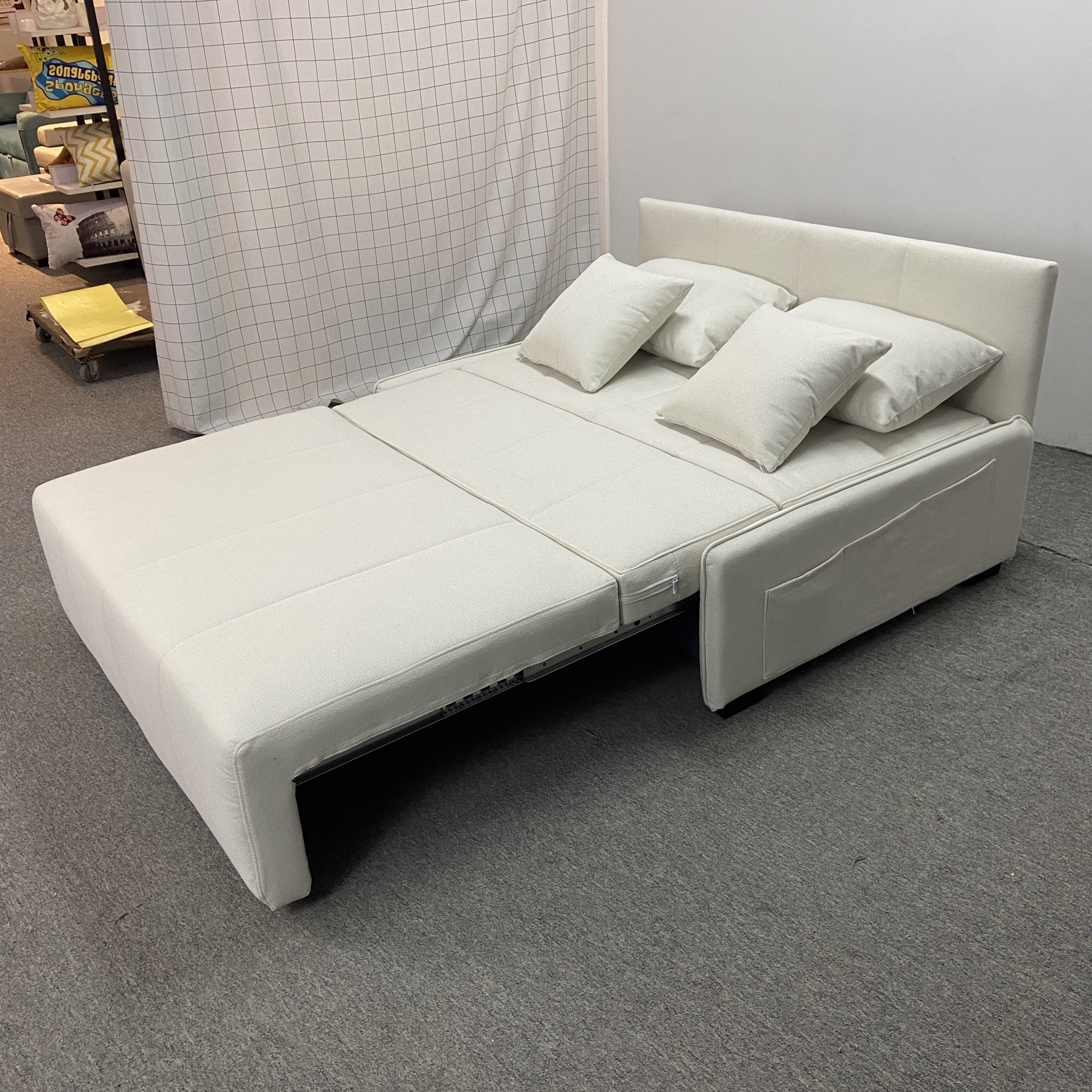 Takumi 6079款矮扶手版本米白色布藝梳化床展開為床的俯視側角，可見床面結構及不銹鋼軌道，背靠向後展開，右扶手可見收納袋，工廠背景