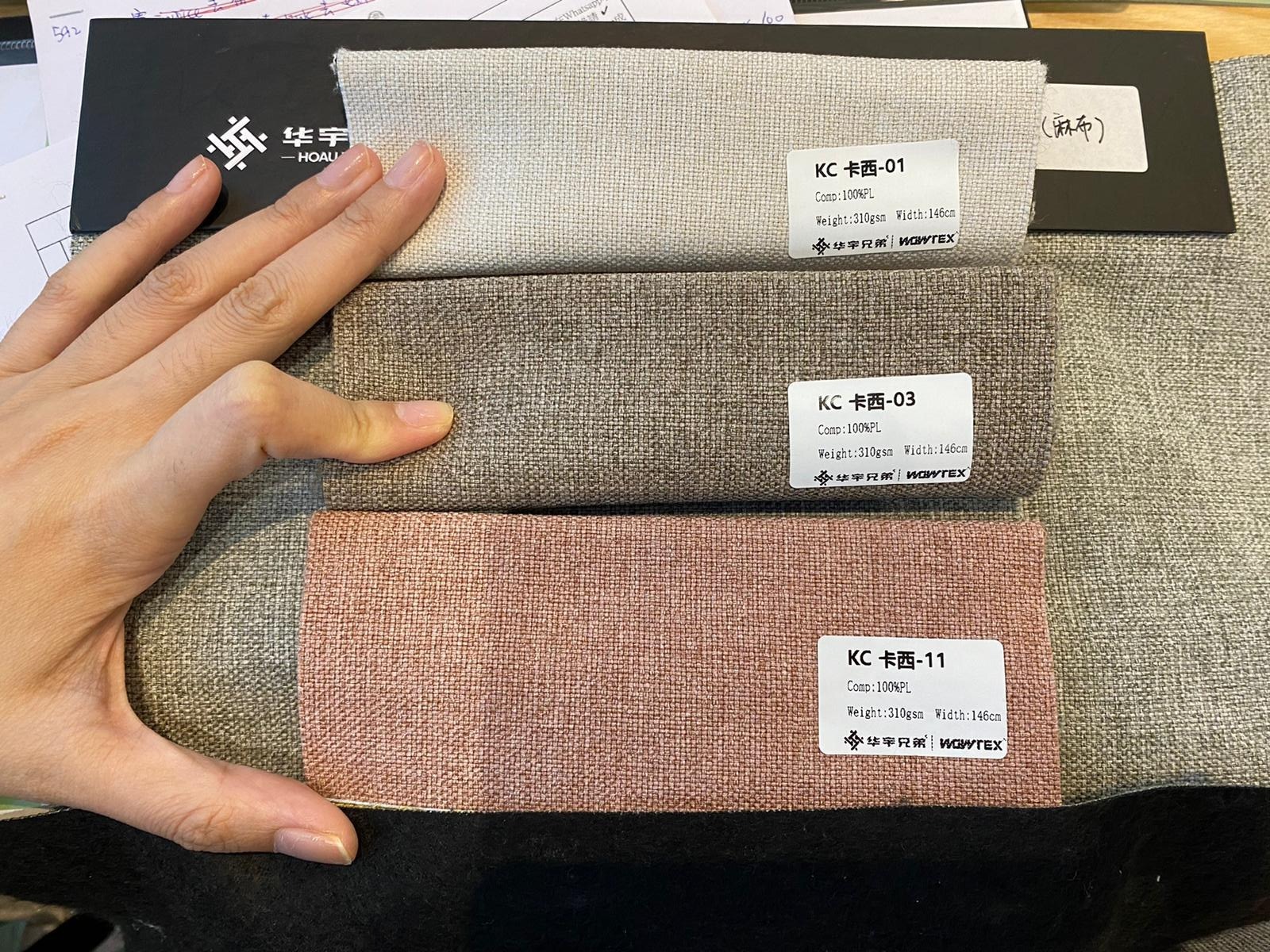 梳化布料色卡三款並排展示，由上至下分別為淺米灰色、炭灰色及粉玫瑰紅色，貼有品牌標籤，一隻手放在旁邊作比例參考