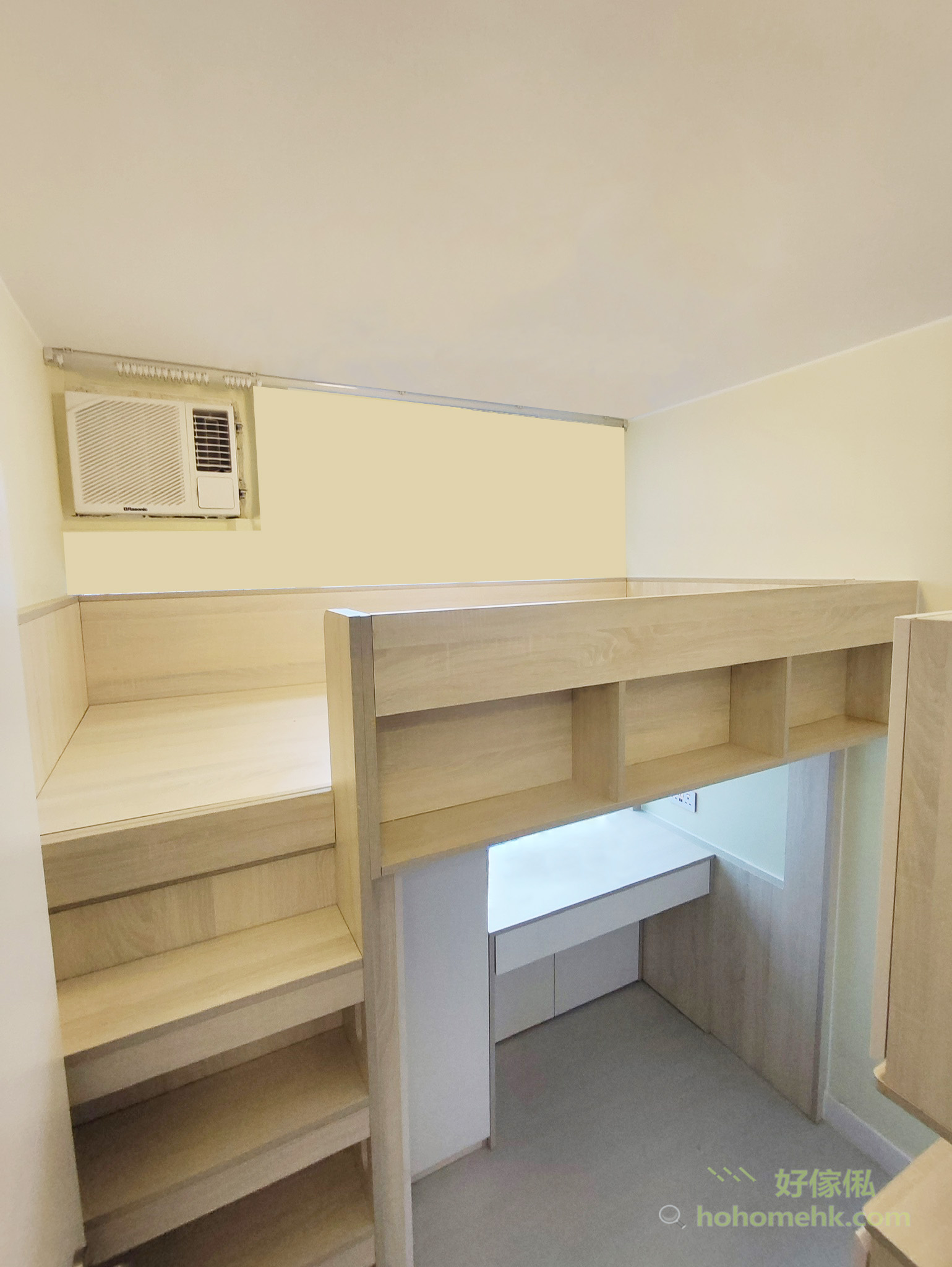 訂造高架床側面角度，淺木色闊踏步梯級由地面向上連接床台，床台側板下方設圍欄書架一排，床台右下方為嵌入式書枱區域配白色桌面及底部收納櫃，書枱區設有燈光，背景為米白色牆身，頂部有窗型冷氣。