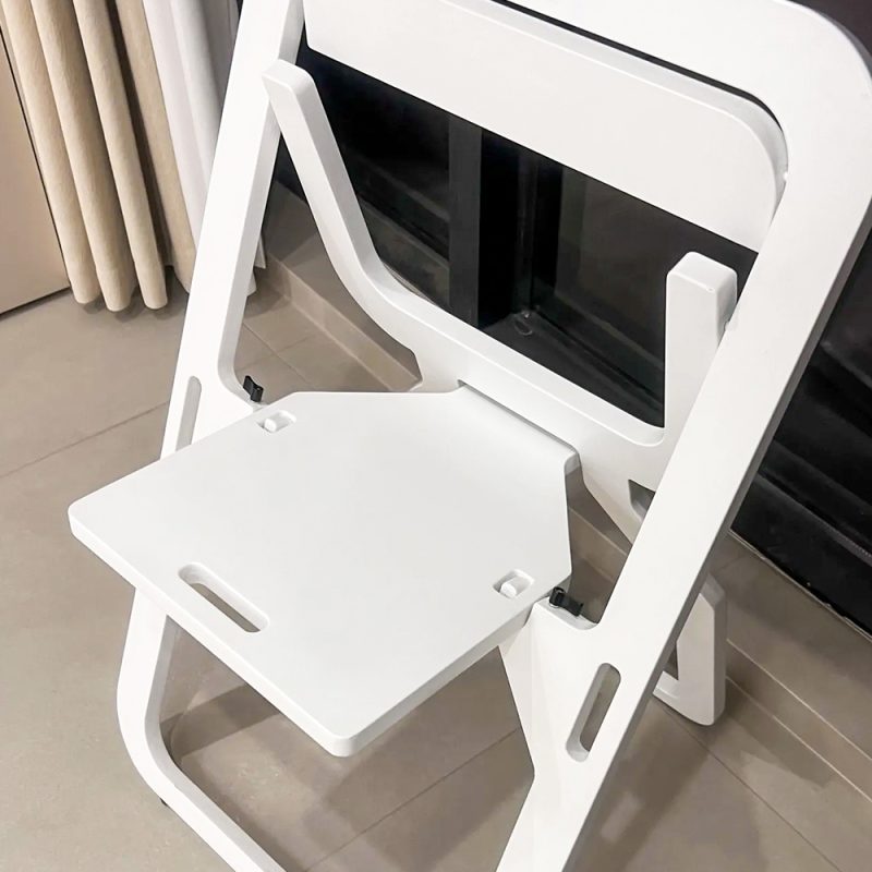 近拍：白色烤漆 Quinn 折疊凳展開狀態，角度由上方俯視，可見方形坐板兩側有提手槽，椅背鏤空矩形設計，黑色金屬鉸鏈固定坐板，背景為淺色地板及玻璃門。