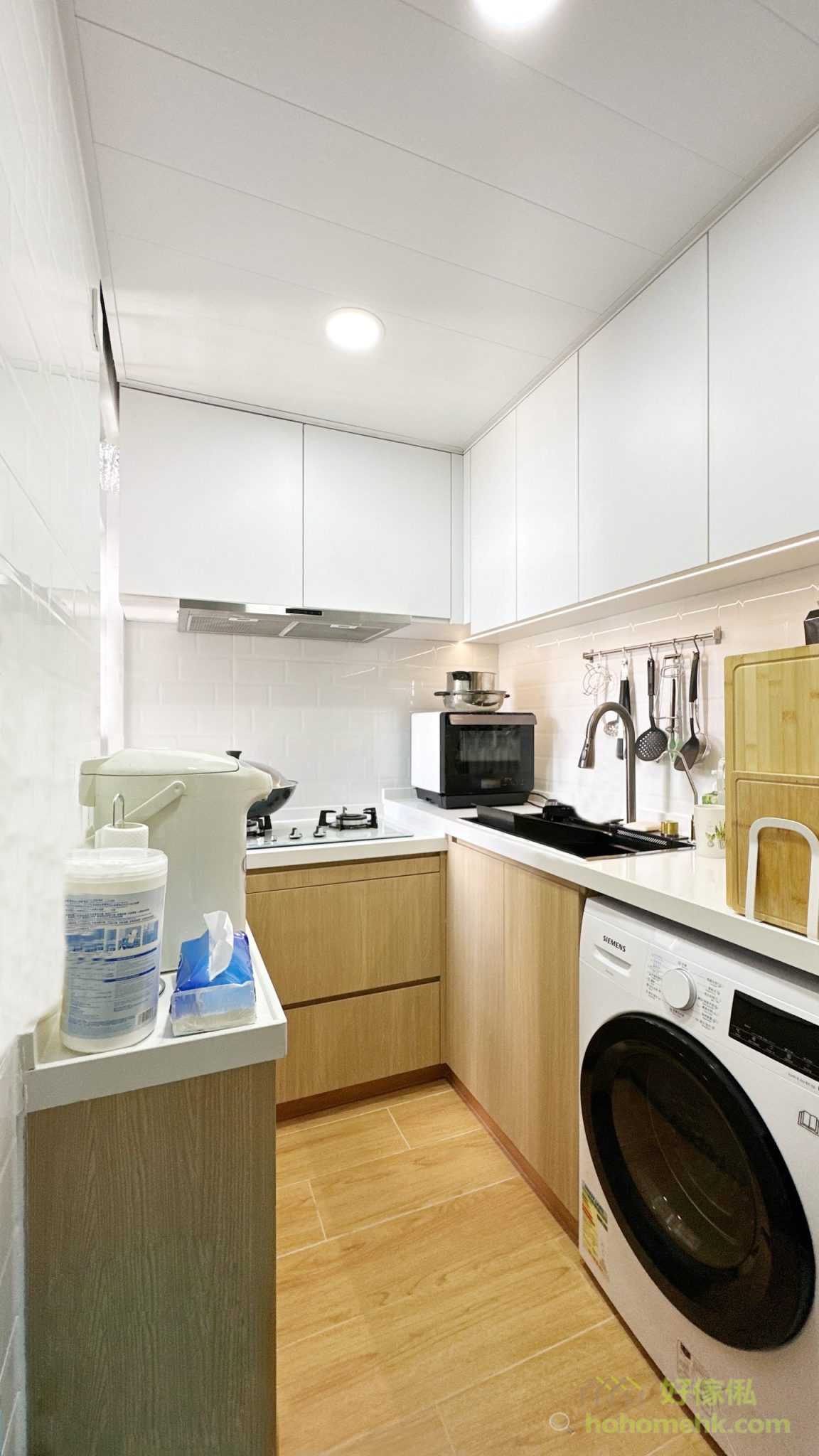 馬鞍山錦駿苑居屋廚房全景，U形廚櫃設計，上層白色無把手吊櫃直頂天花，下層原木紋大抽屜地櫃，白色石英石工作枱面，鋅盤旁嵌入洗衣機，白色地磚配木紋牆磚，空間通透整潔