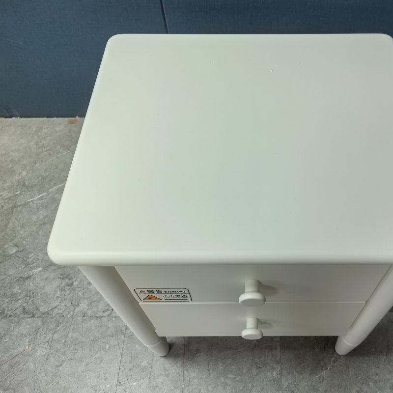 由上斜望落嘅實拍圖，Carey床頭櫃奶油白檯面呈方形，四角採用大弧度圓角處理，枱邊順住向下收落抽屜面，抽屜面貼有警告標貼，底部見到灰色石紋地面同深藍牆身。