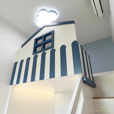 訂造閣樓床上層屋仔造型特寫仰拍，白色板材頂部以深藍色斜屋頂形狀收邊，正面嵌入深藍色格形窗框，四格窗裝飾造型立體；圍欄為藍白間條垂直條板設計；上方為雲朵造型白色吸頂燈；房間天花及牆身為白色