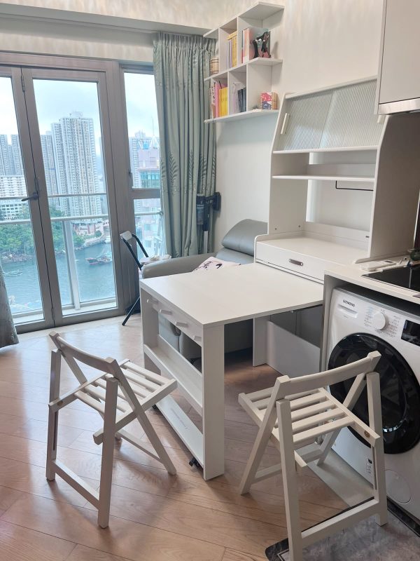 香港細單位廚房位置，Nova白色實木伸縮餐枱完全拉開至長方形枱面，兩張白色木製摺椅斜靠喺枱邊，右邊係白色多層餐邊櫃連玻璃門格同抽屜，左邊落地玻璃窗望出海景陽台，地下係淺色木地板，西門子洗衣機貼位擺放。