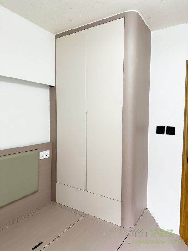 主人房的設計重點在於因應套廁門口的位置，特別訂造了斜角地台，避免出入廁所時的不便。此外，卡其色的地台床與圓邊衣櫃相互呼應，不僅提升了安全性，還增添了居室的溫馨感。床頭綠色軟墊的設計，更為整體空間帶來了一抹柔和的色彩。