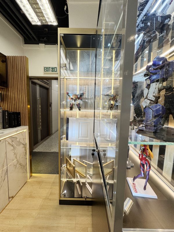 門市走廊旁擺放一座八呎高窄身玻璃展示櫃，香檳金色鋁框配透明玻璃門及多層玻璃層板，櫃內燈帶照亮機械人模型，旁邊連接另一座黑框收藏櫃。
