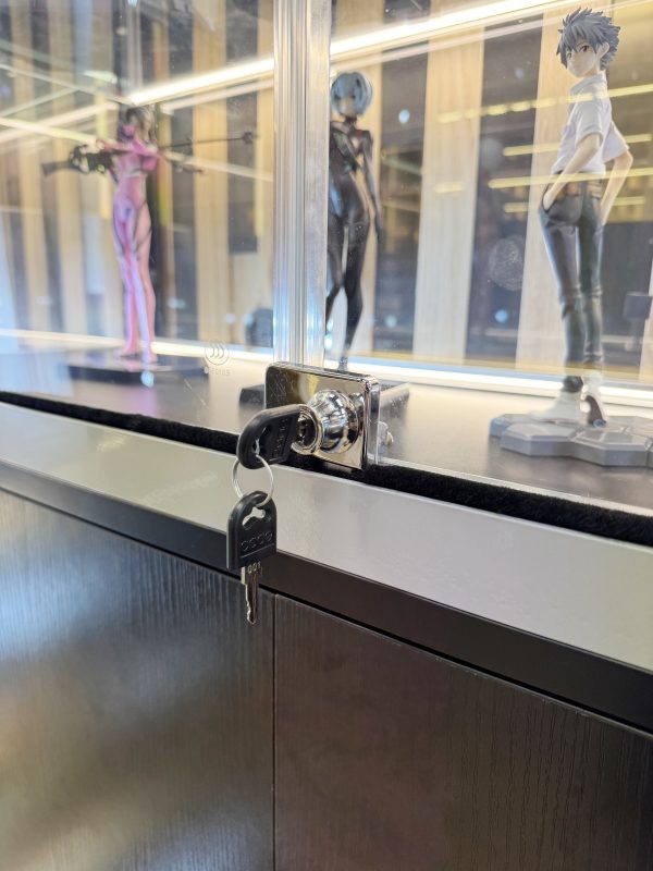 玻璃展示櫃趟門近鏡，兩塊透明玻璃交疊位置裝有銀色金屬鎖，黑色鎖匙插在鎖芯上，後方層板陳列粉紅、黑色及白色人偶模型。