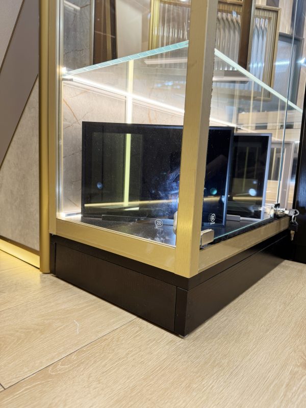 香檳金色鋁框玻璃展示櫃底部近鏡，透明玻璃門內放有黑色板件，櫃身承托於內縮黑色木腳座上，腳座沿淺木色地板靠近牆邊擺放。