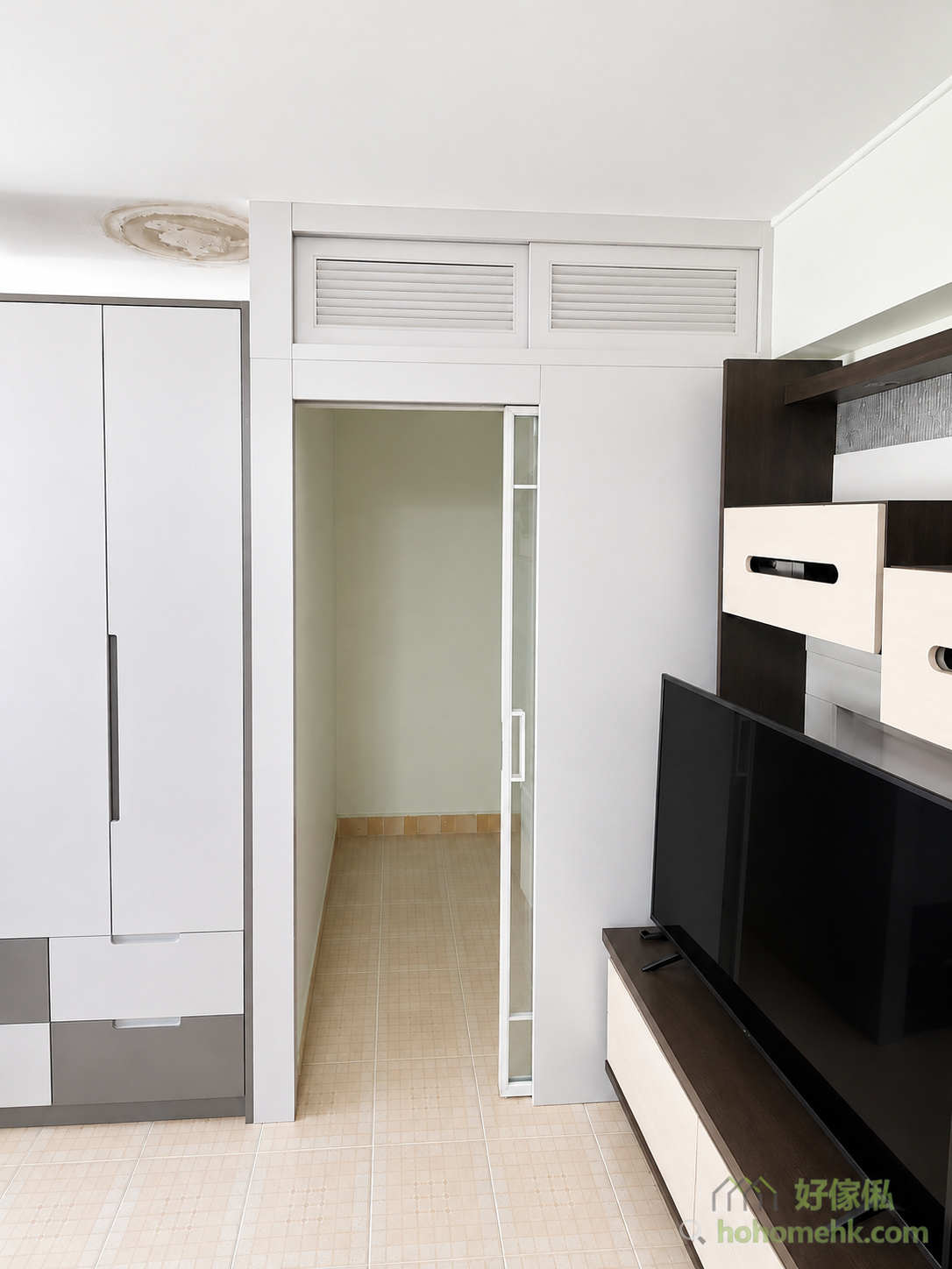 白色公屋客廳以間房牆劃出獨立空間，入口設磨砂玻璃趟門，門頂配兩扇百葉氣窗，左側貼近白灰色衣櫃，右側為黑白電視櫃。