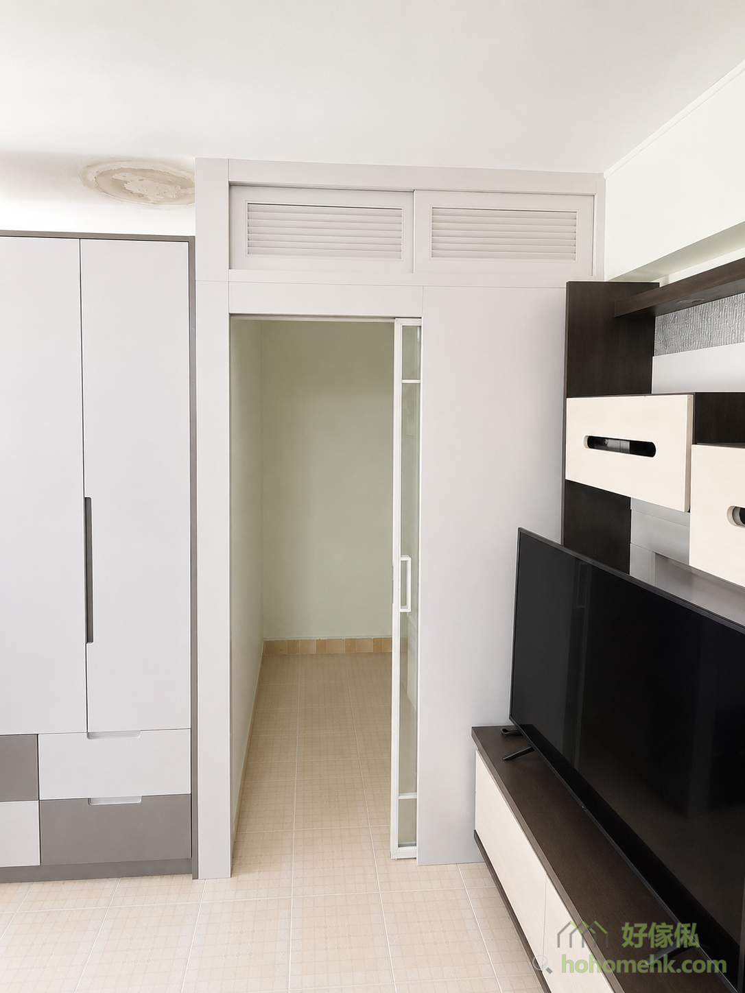 白色公屋客廳以間房牆劃出獨立空間，入口設磨砂玻璃趟門，門頂配兩扇百葉氣窗，左側貼近白灰色衣櫃，右側為黑白電視櫃。
