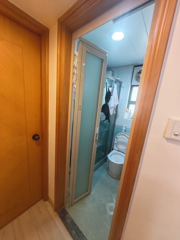 杏花邨住宅走廊望入浴室，銀色金屬框PD趟摺門向左摺開，門扇沿原有木色門框收起；室內可見白色坐廁、灰色牆地磚、晾掛衣物及窗戶。
