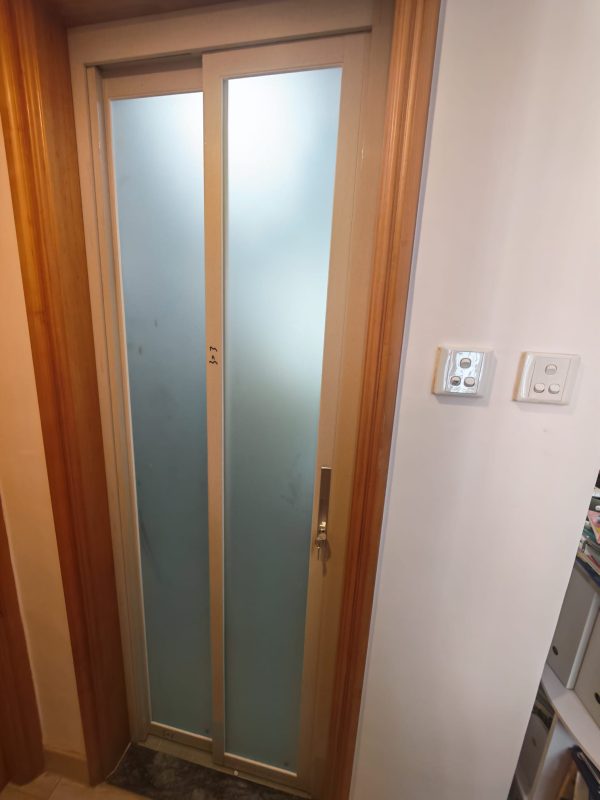 正面近拍杏花邨浴室關閉狀態嘅PD趟摺門，銀色金屬框包住兩幅藍灰色半透明磨砂門板，門身嵌於原有木色門框，右邊白牆設有兩組開關。