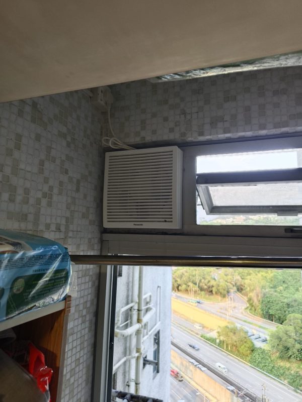 灰白色細格仔磚牆旁嘅鋁窗上方，左側裝有白色 Panasonic 方形百葉面抽氣扇，右側窄窗向外打開，窗外可見樓下道路及樹木。