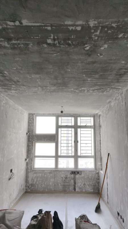 狹長客廳的灰色天花及兩側牆身已大範圍剷底，盡頭白色鋁窗保留防盜花，地面覆蓋白色保護物料，右牆斜放一把長柄掃把。