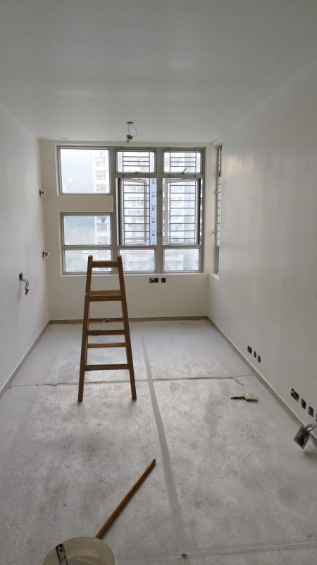 狹長客廳完成白色牆身及天花油漆，盡頭設有多格鋁窗及防盜窗花，灰色地面鋪上保護物料，室內放置木梯及施工掃把。