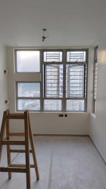 客廳窗邊牆身完成批灰油漆，正面及右側設有多格鋁窗與防盜窗花，白色牆面配灰色保護地面，左前方放置木梯。