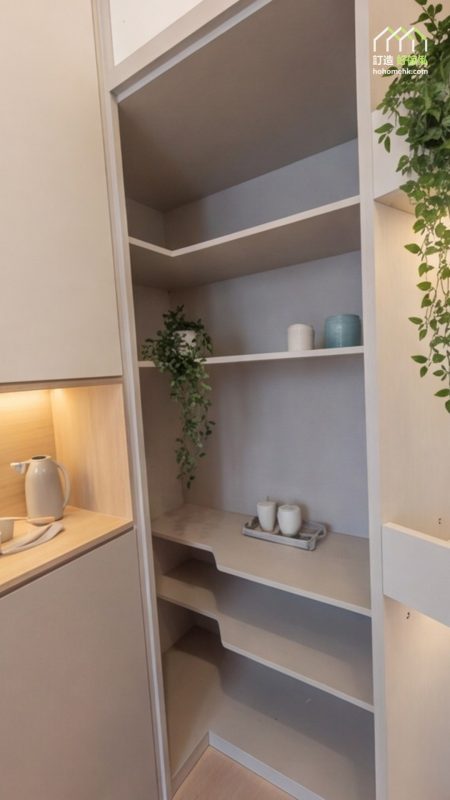 灰色開放展示櫃設多層層板，淺色櫃框包圍灰調背板，架上擺有杯具、花器及垂吊綠植，旁邊木紋中空枱面透出暖光。