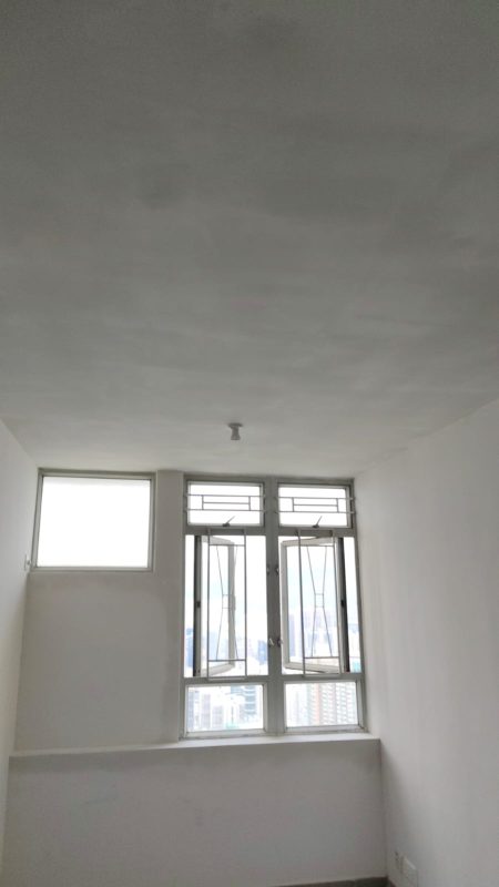 狹長睡房兩側為白色牆身，天花保留灰白色批灰紋理，盡頭設雙扇鋁窗及兩個高位玻璃窗，窗台下方牆身已修平，地面為灰色施工面。